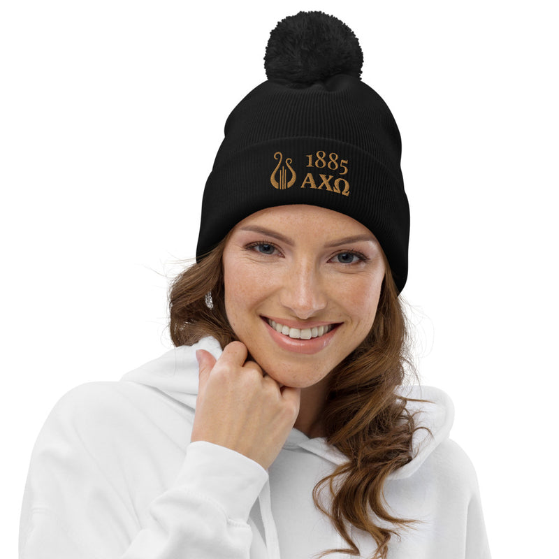 Alpha Chi Omega 1885 Lyre Pom Pom Beanie shown in black with gold embroidery on model
