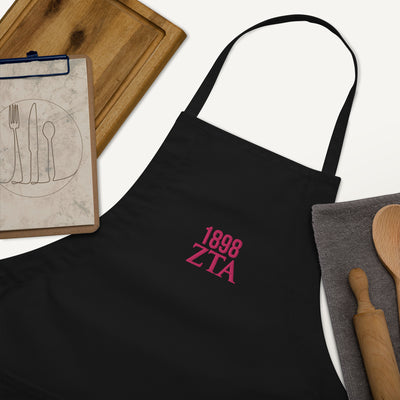 Zeta Tau Alpha 1898 Founding Year Embroidered Apron in black with pink thread shown with kitchen utensils