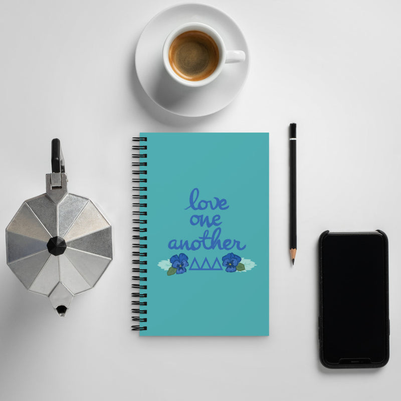 Tri Delta Love One Another Spiral Notebook shown with coffee